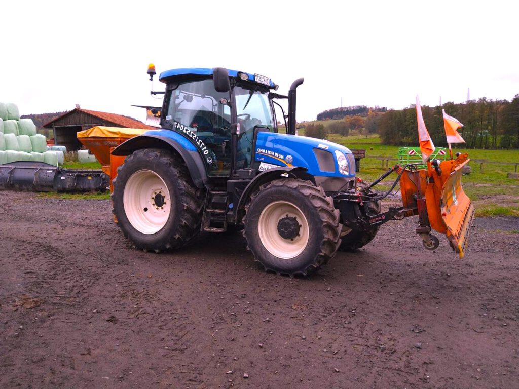 New Holland Traktor trägt einen Schneepflug vor sich.