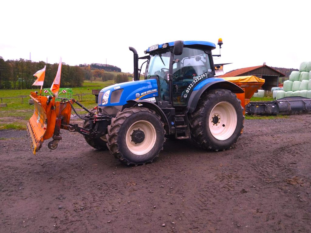 New Holland Traktor trägt einen Schneepflug vor sich.