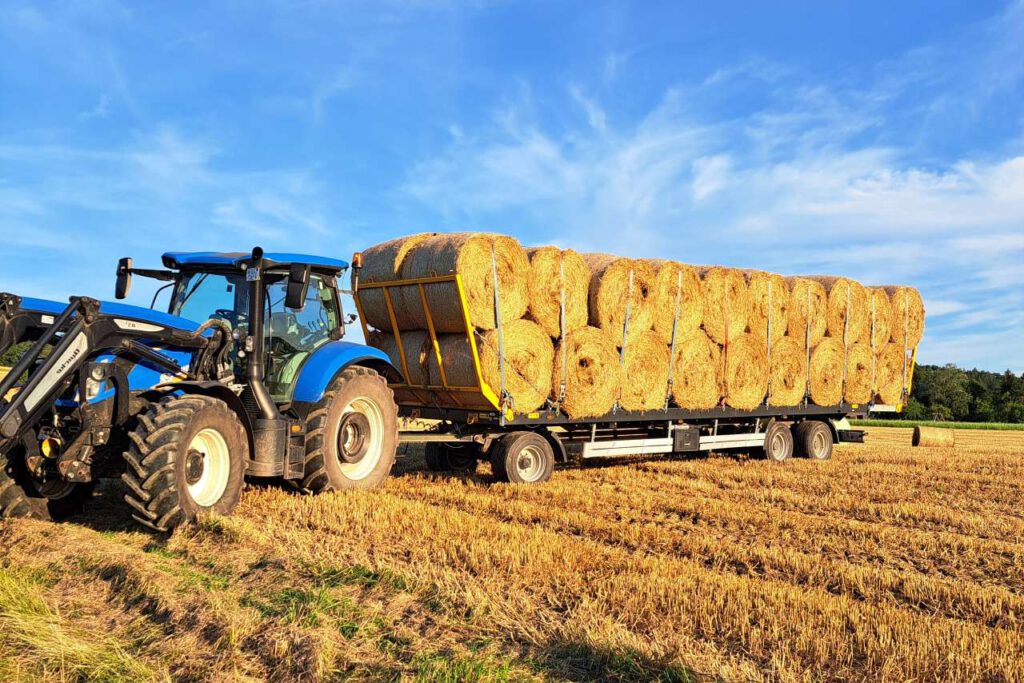 New Holland Traktor T7.210 Transportarbeiten von Stroh Rundballen auf einem Auflieger, der durch ein Stoppelfeld gezogen wird.